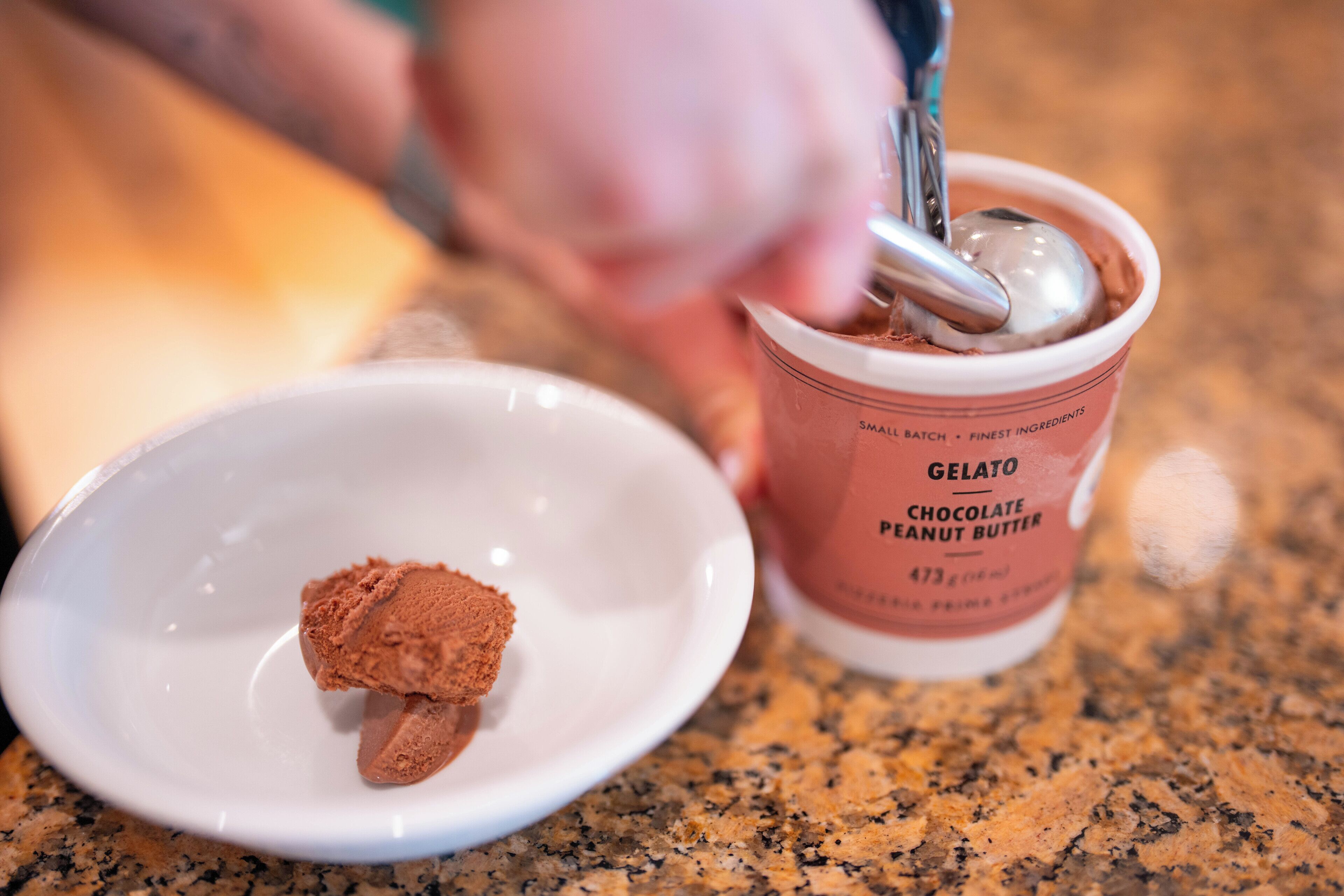 A scoop of chocolate peanut butter gelato in a branded cup and a small scoop of the same gelato on a plate.