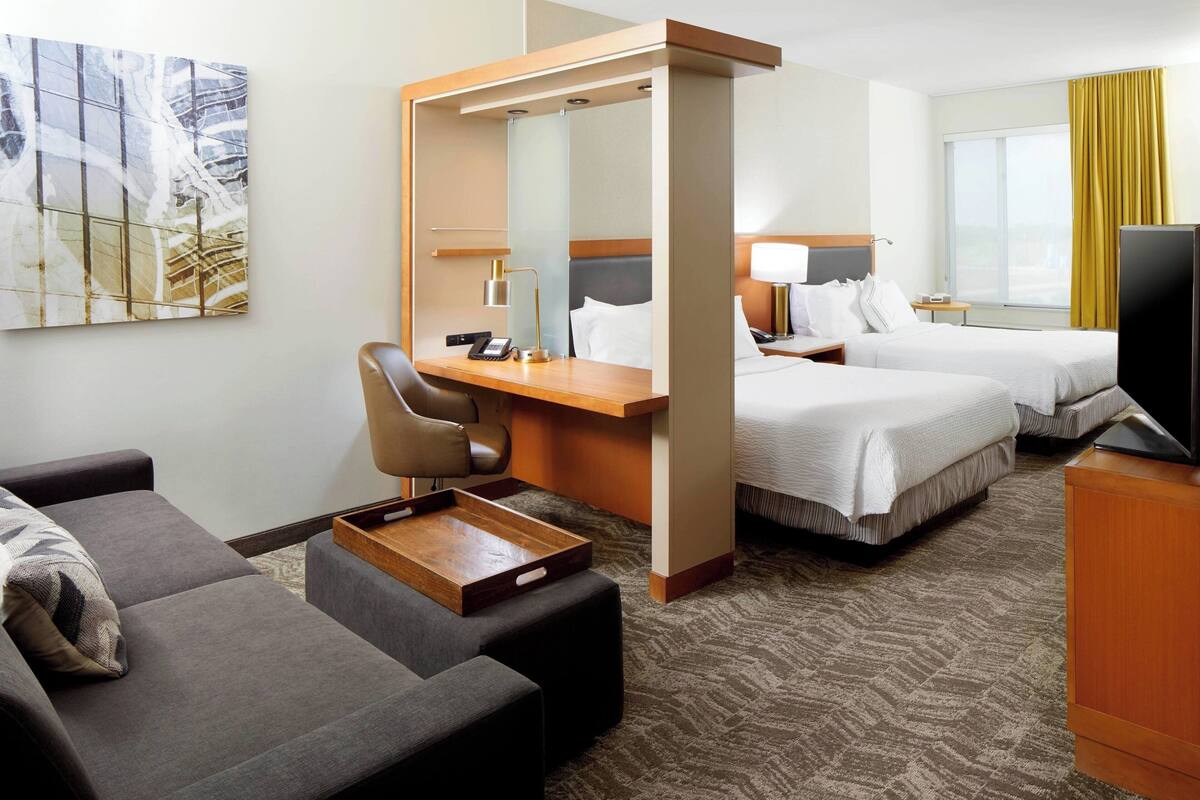 A hotel room with a desk, two beds, a sofa, a TV, and a large window with curtains.