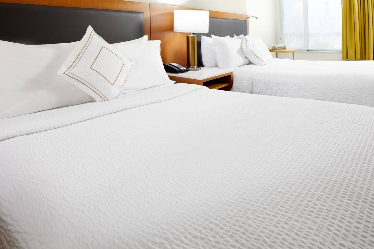 A hotel room with two beds, a bedside table with a lamp, and a window with curtains.