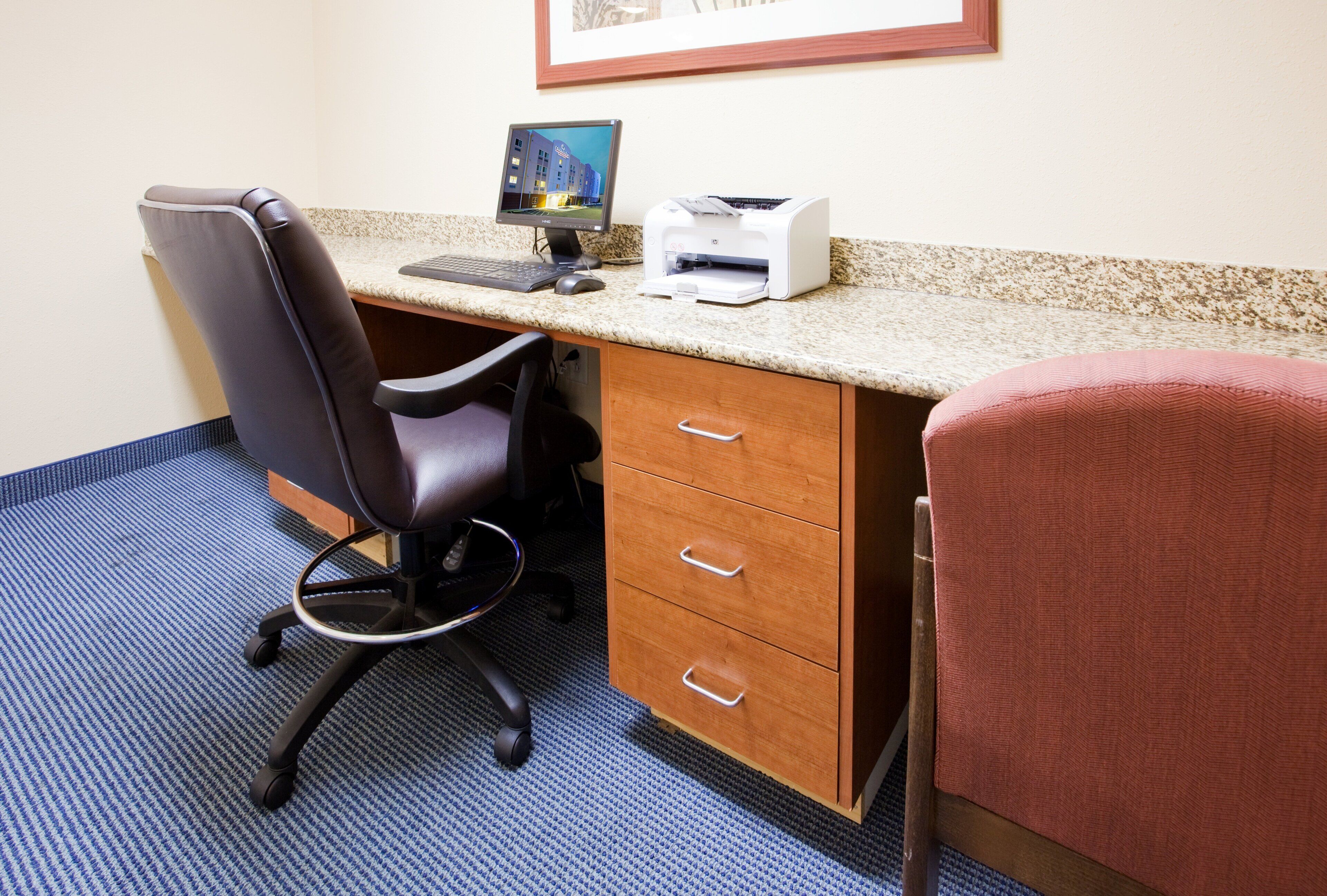 A hotel room with a desk, chair, computer, printer, and a bed with a red blanket.
