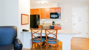 A compact kitchen with wooden cabinets, a black refrigerator, a microwave, and a coffee maker. There are two wooden chairs with blue cushions. - Candlewood Suites Jacksonville by IHG (Jacksonville)