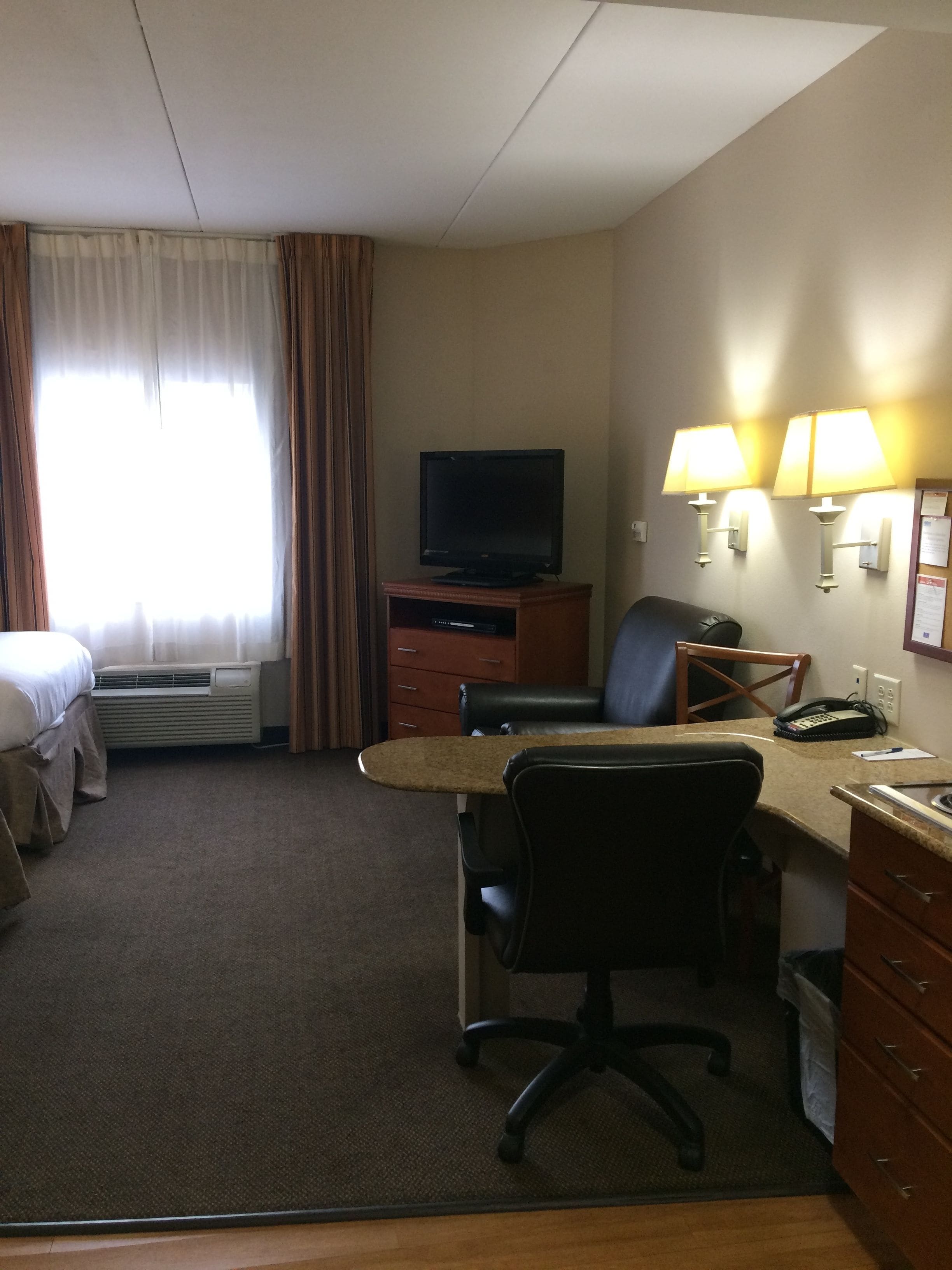 Ett hotellrum med en säng, ett skrivbord med en stol, en tv, en telefon och en lampa.