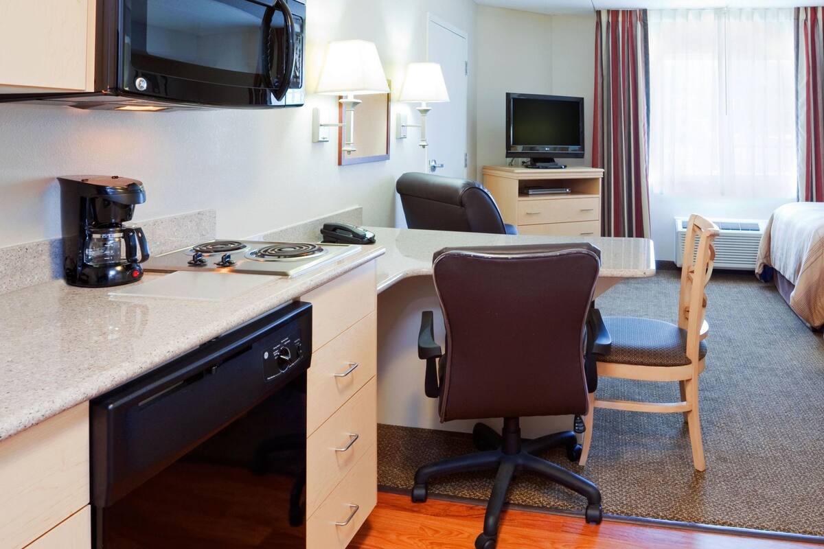 A hotel room with a kitchenette, a desk with a chair, a bed, a TV, and a window with curtains.