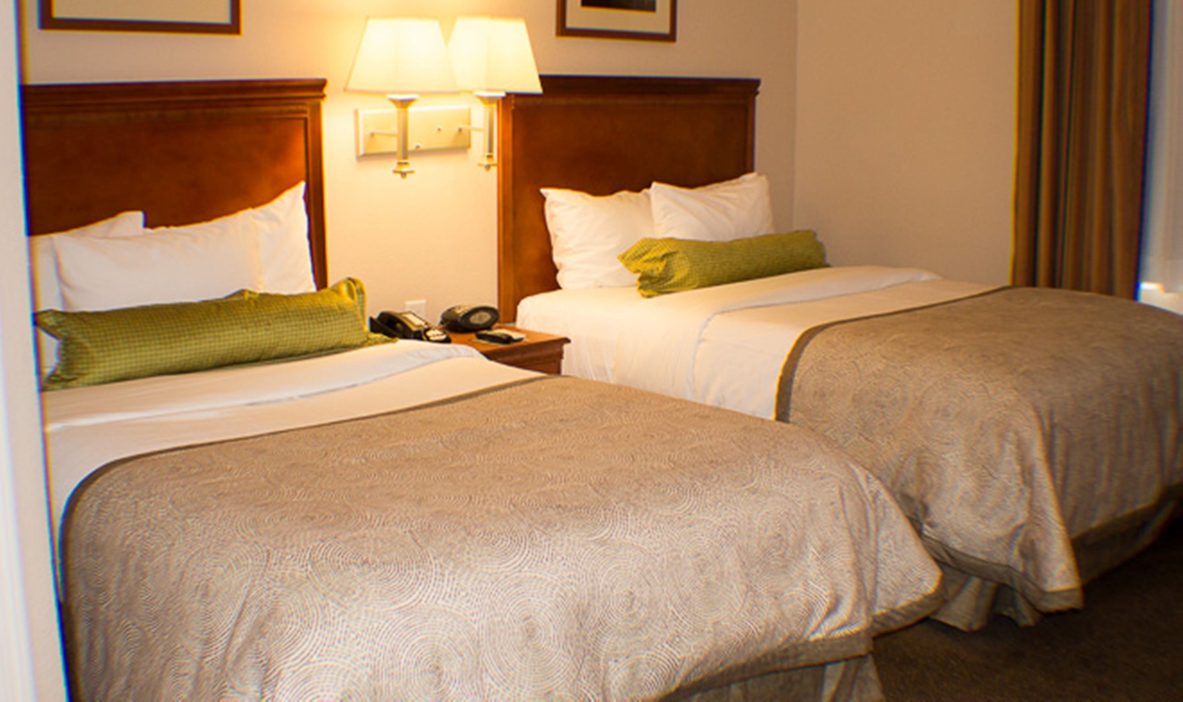 A hotel room with two beds, each with a wooden headboard, white bedding, and a green throw pillow. There is a nightstand with a lamp and a telephone.