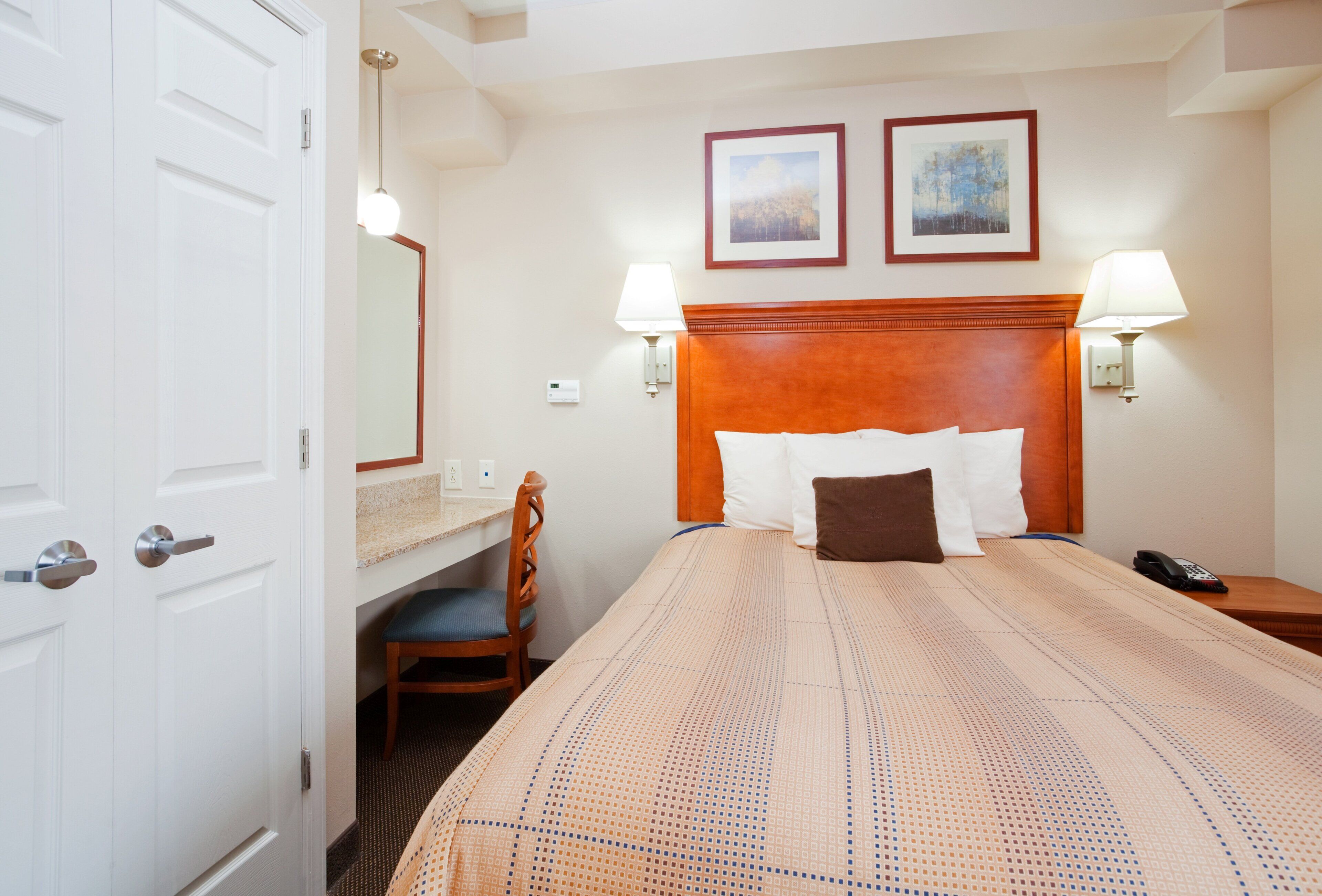 A hotel room with a bed, bedside lamps, a desk with a chair, a telephone, and two framed pictures on the wall.