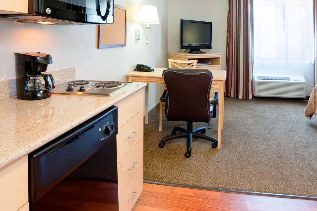 A hotel room with a kitchenette, a desk with a computer, a TV, a bed, and a window with curtains.