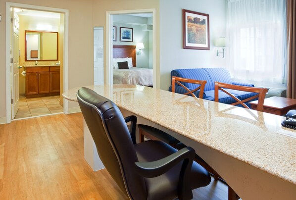 A hotel room with a kitchen area, a dining table with chairs, a bed with a headboard, a blue sofa, and a window with curtains. - Candlewood Suites Jacksonville by IHG (Jacksonville)