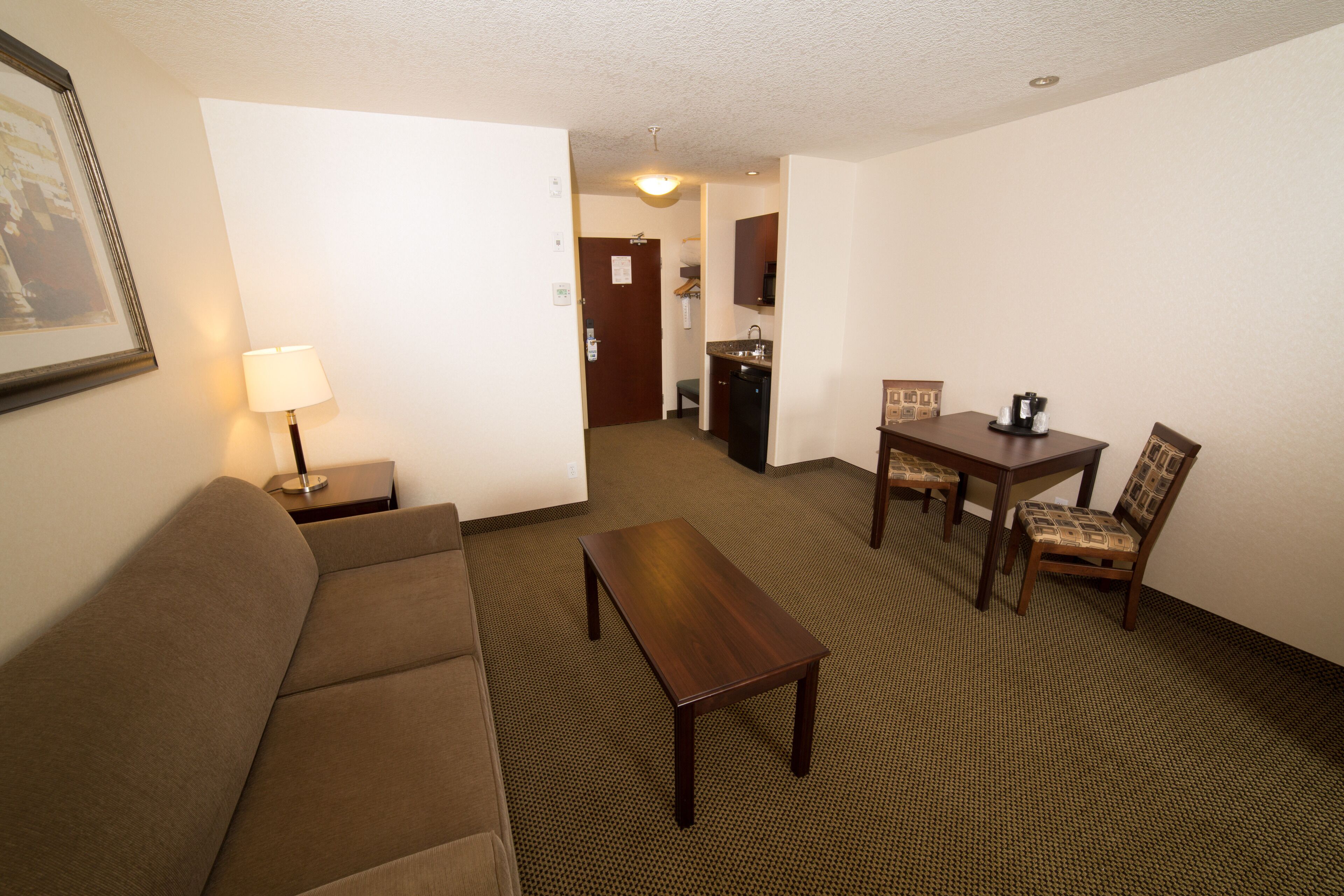 A hotel room with a sofa, a wooden coffee table, a dining table with chairs, a small kitchen area, and a framed picture on the wall.