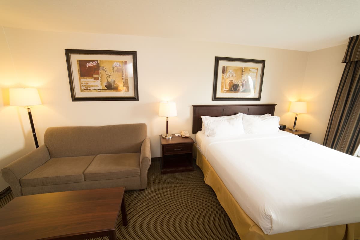 A hotel room with a bed, a sofa, a nightstand with a lamp, and two framed pictures on the wall.