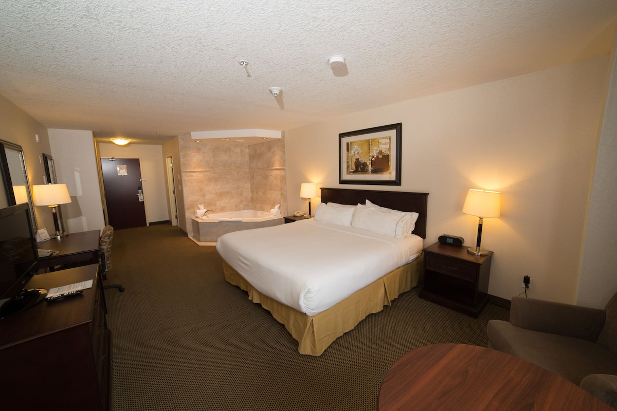 A hotel room with a large bed, a desk with a chair, a small table, a sofa, a painting on the wall, and a bathroom with a bathtub and shower.