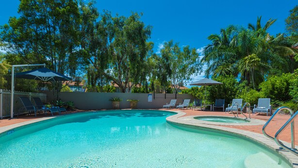 A swimming pool with clear blue water, surrounded by lounge chairs and umbrellas, with trees and a fence in the background. - At The Sound (Noosaville)