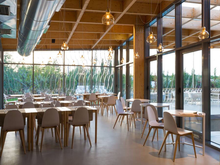 A spacious dining area with wooden tables and chairs, large windows offering a view of greenery, and hanging pendant lights.