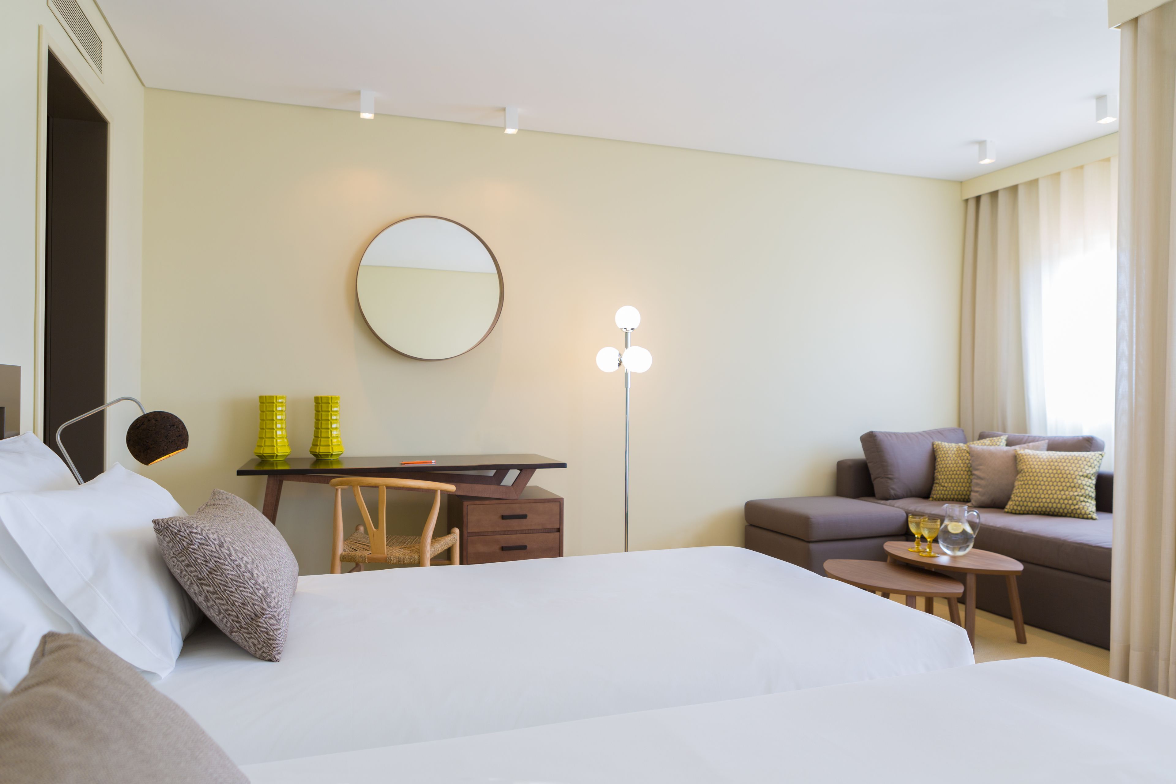 Quarto de hotel com cama, escrivaninha, cadeira, sofá, espelho redondo e mesa pequena com luminária.
