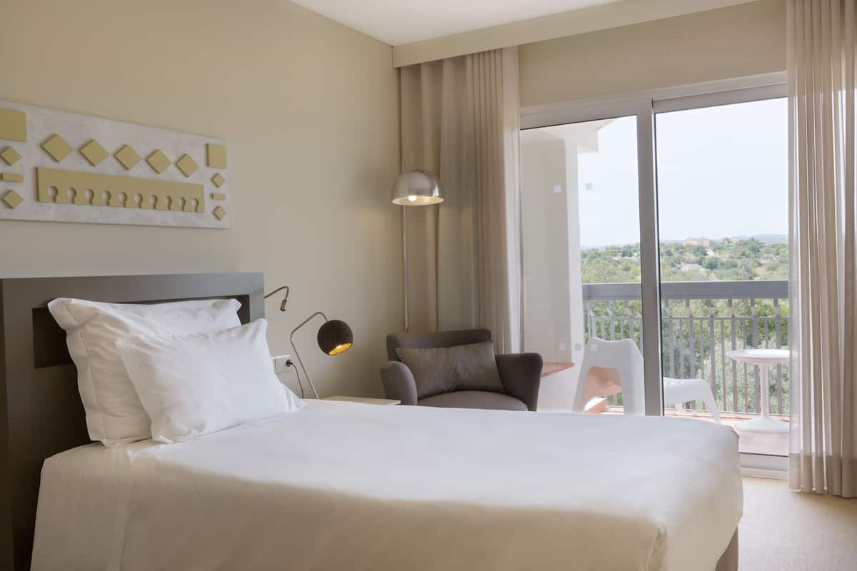 A hotel room with a large bed, a chair, a desk, a lamp, and a balcony with a view.