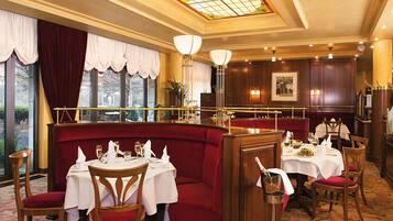Een restaurant met rode bekleden zitmogelijkheden, ronde tafels met witte tafellakens en een kroonluchter.