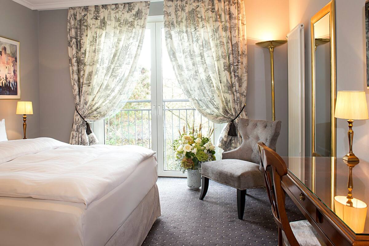 A hotel room with a large bed, a chair, a lamp, a mirror, and a window with curtains.