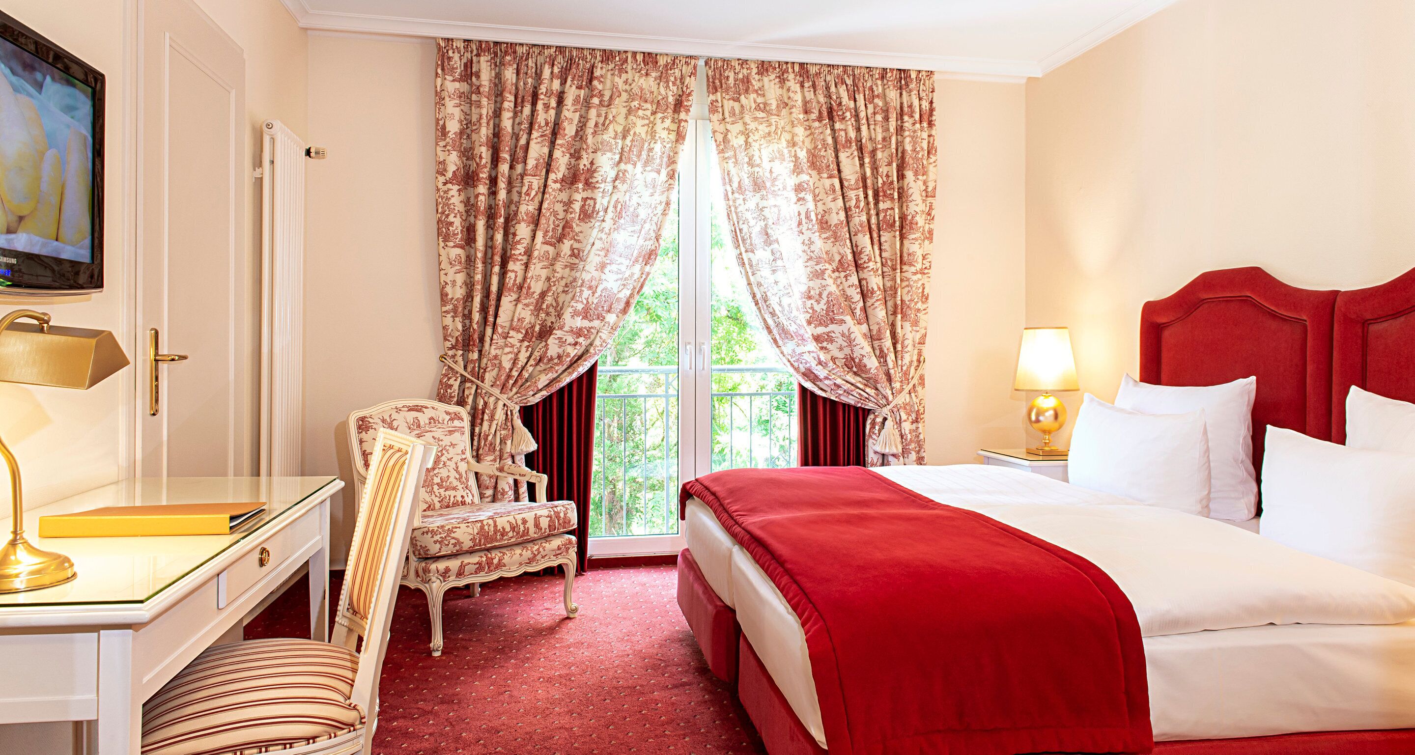 A hotel room with a bed, a desk with a lamp, a chair, and a large window with curtains.
