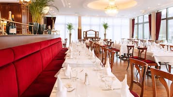 A restaurant with a long dining table set for a meal, red upholstered seating, and large windows.