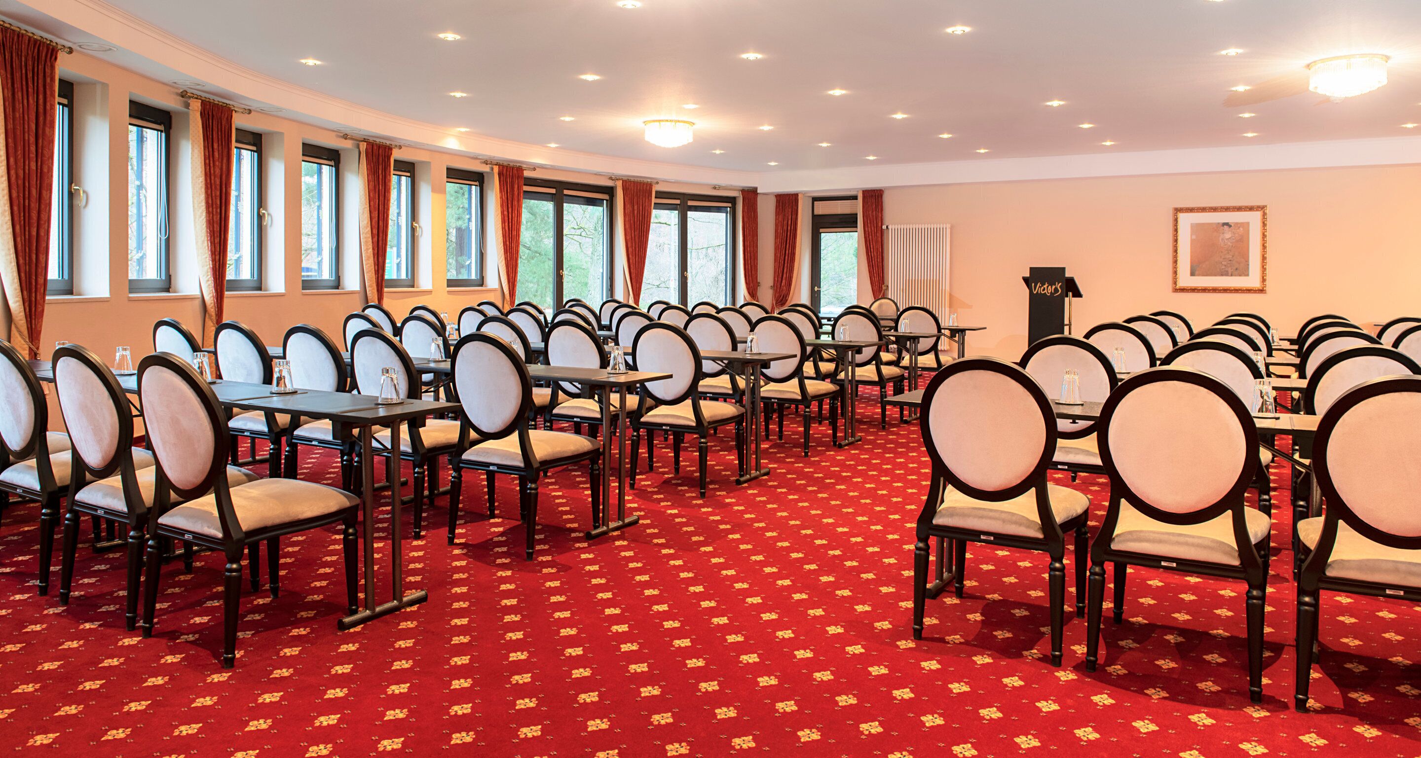 Une salle de conférence avec des rangées de tables et de chaises, de grandes fenêtres avec des rideaux et un sol recouvert d’une moquette rouge.