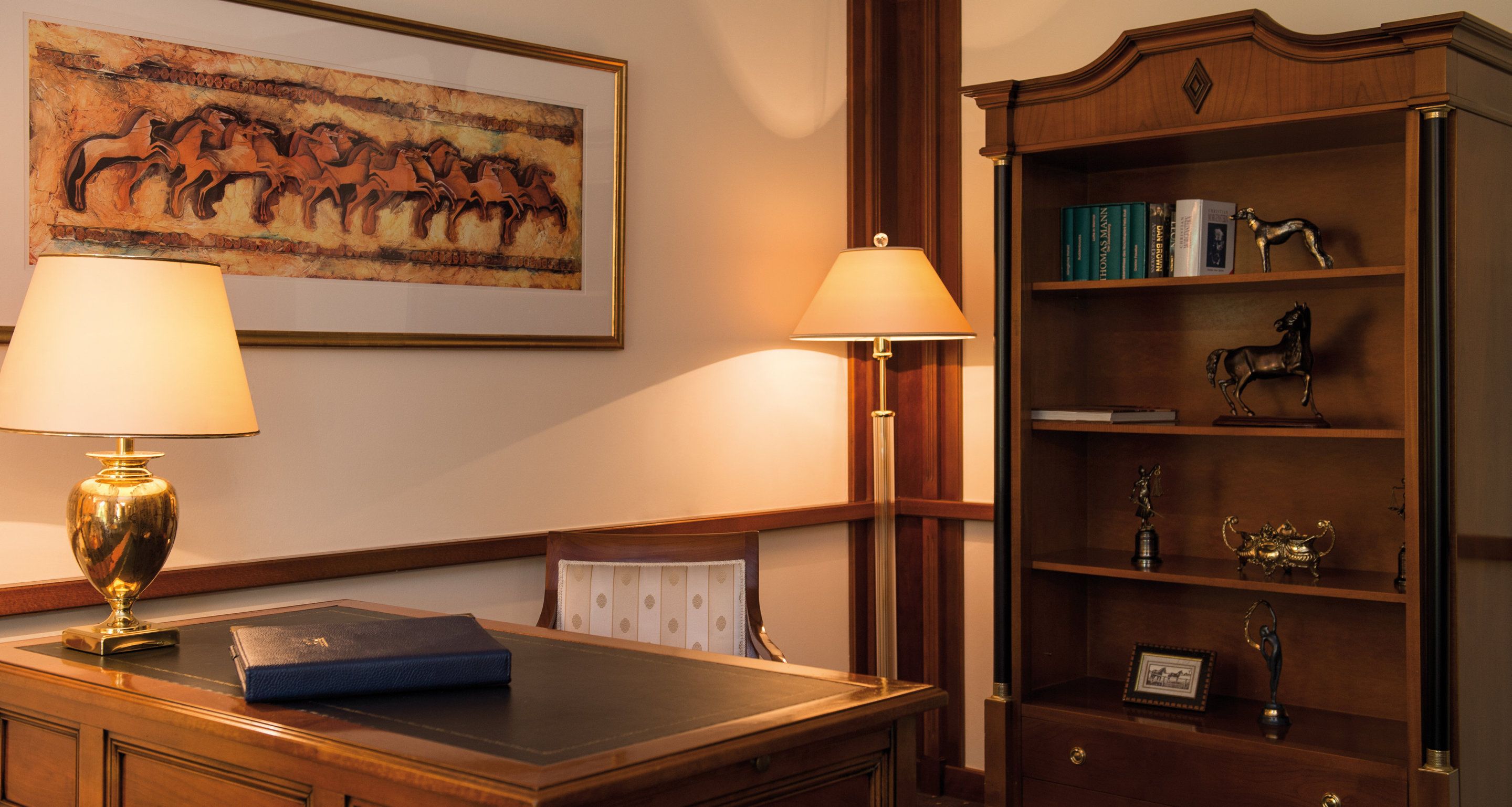 Una sala de estudio con un escritorio, dos lámparas, una estantería con libros y figuritas de caballos, y un cuadro de caballos en la pared.