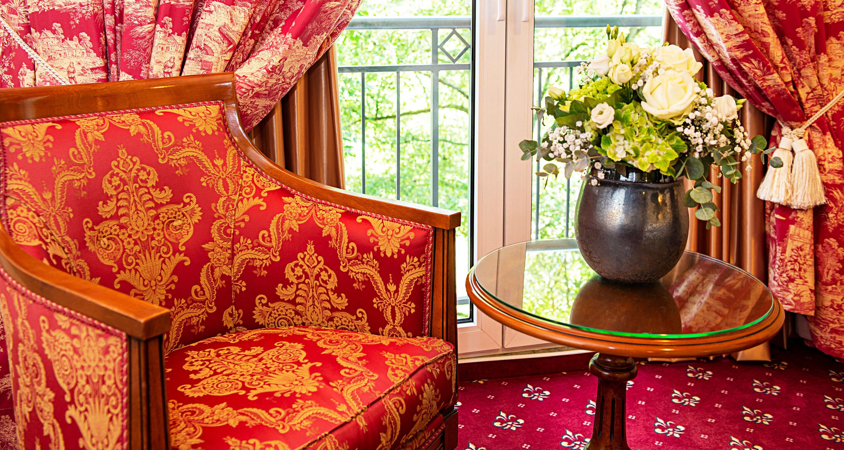 Una butaca tapizada de rojo con un diseño dorado, una mesa de vidrio con un jarrón de flores y vista a zonas verdes a través de una ventana.