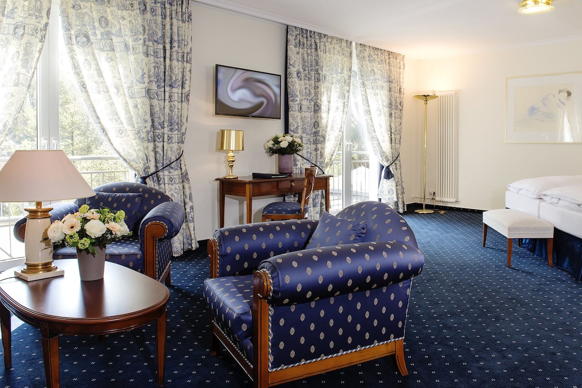 A hotel room with a bed, two armchairs, a small table with a lamp, a vase of flowers, and a painting on the wall.