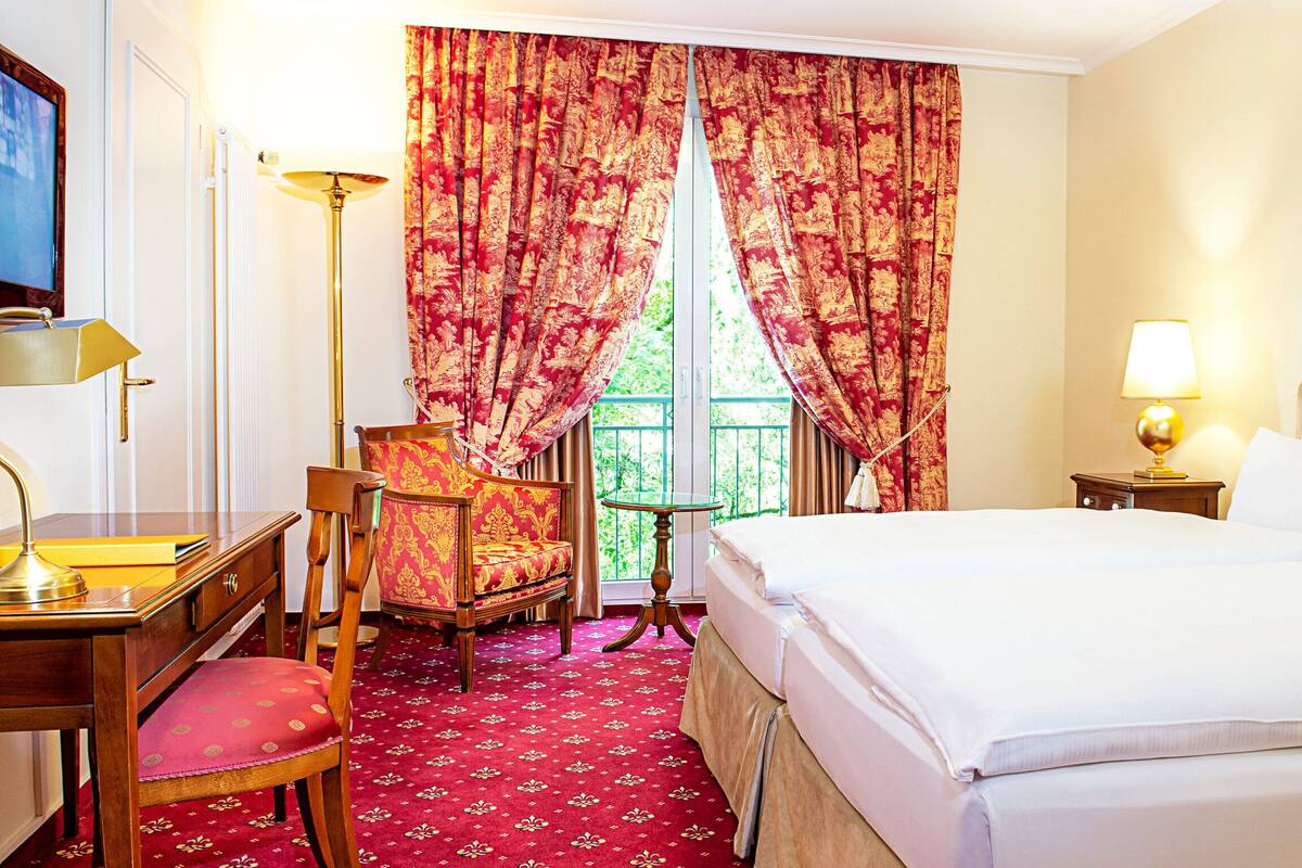 A hotel room with a bed, a desk with a lamp, a chair, a small table, and curtains.