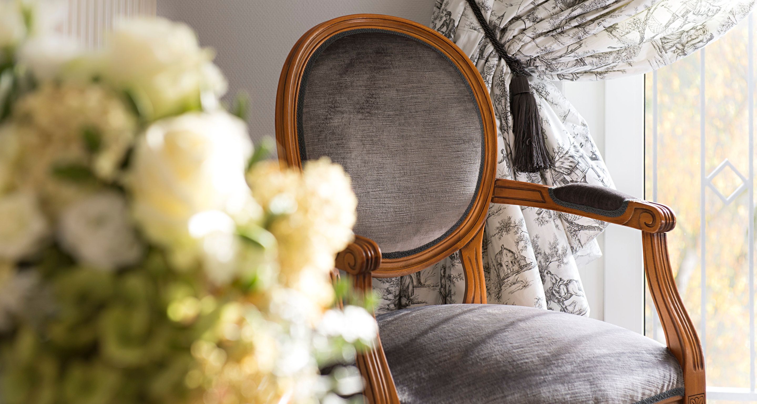A wooden chair with a grey upholstered seat and backrest, positioned near a window with patterned curtains.