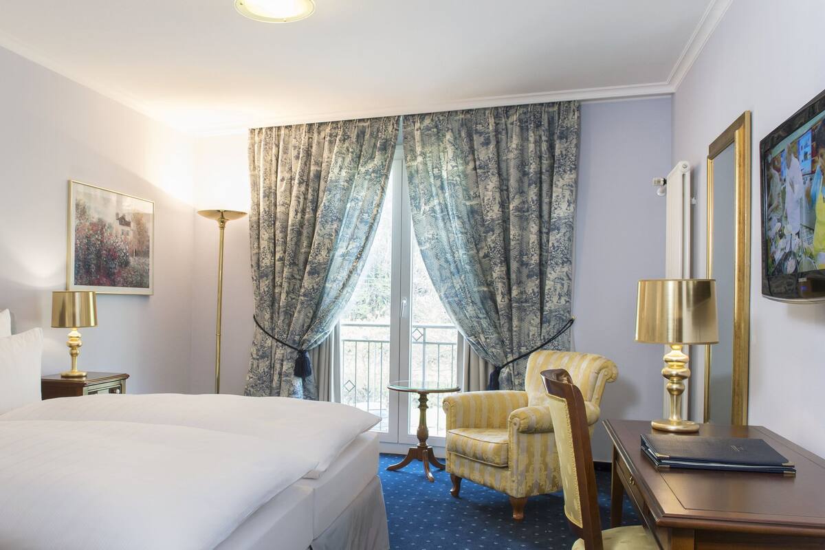 A hotel room with a large bed, a chair, a desk, and a window with curtains.