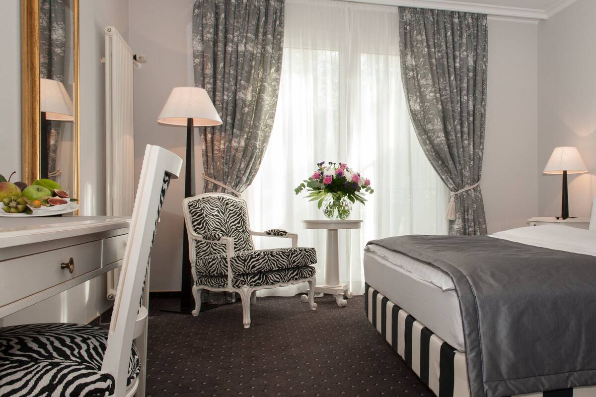 A hotel room with a bed, a chair, a small table with a lamp, a mirror, and a window with curtains.