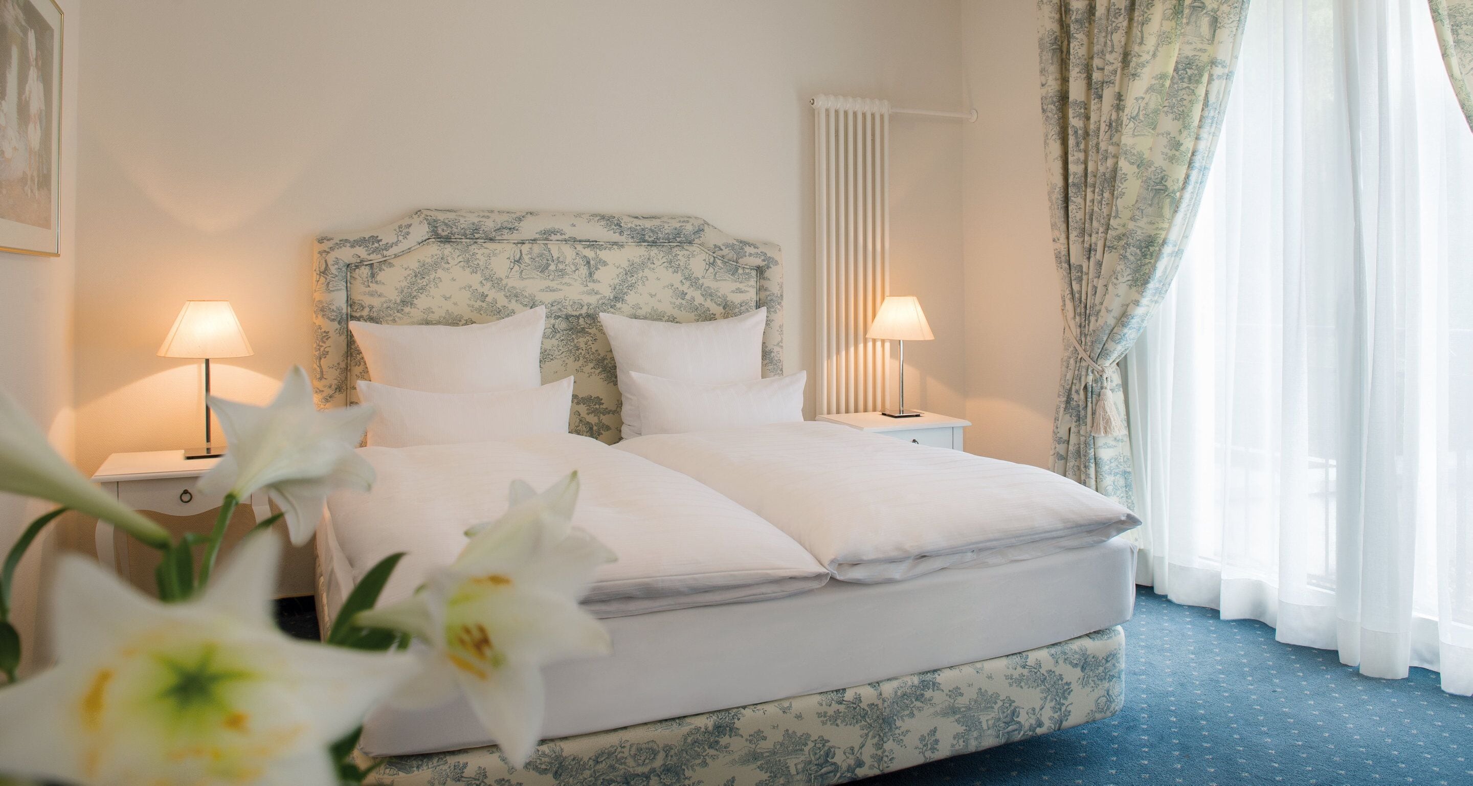 A neatly made bed with white linens and a floral-patterned headboard, two bedside lamps, and a window with sheer curtains.