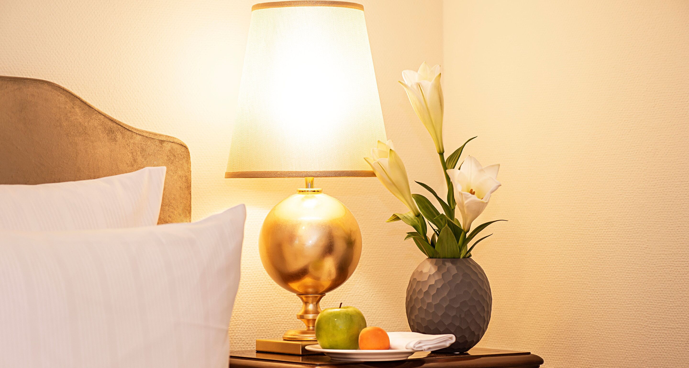 A neatly made bed with white linens, a bedside lamp, a vase with white flowers, and a plate with an apple and a banana.