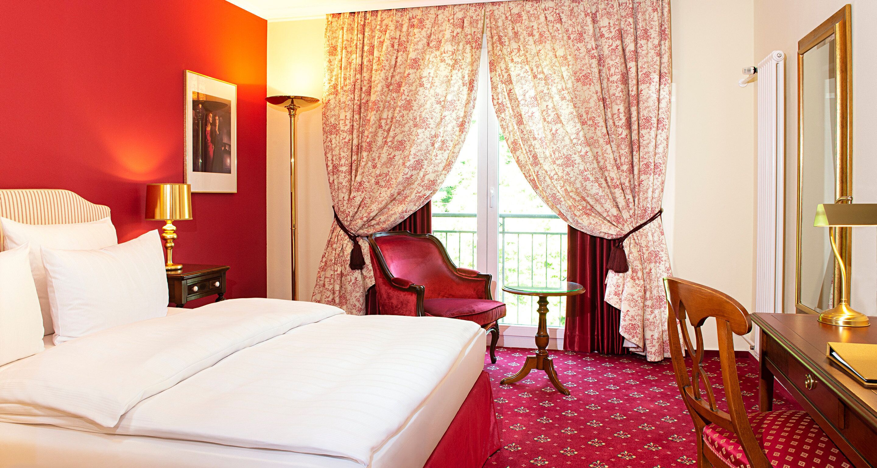 Ein Hotelzimmer mit einem großen Bett, einem roten Sessel, einem kleinen Tisch und einem Schreibtisch mit einer Lampe.
