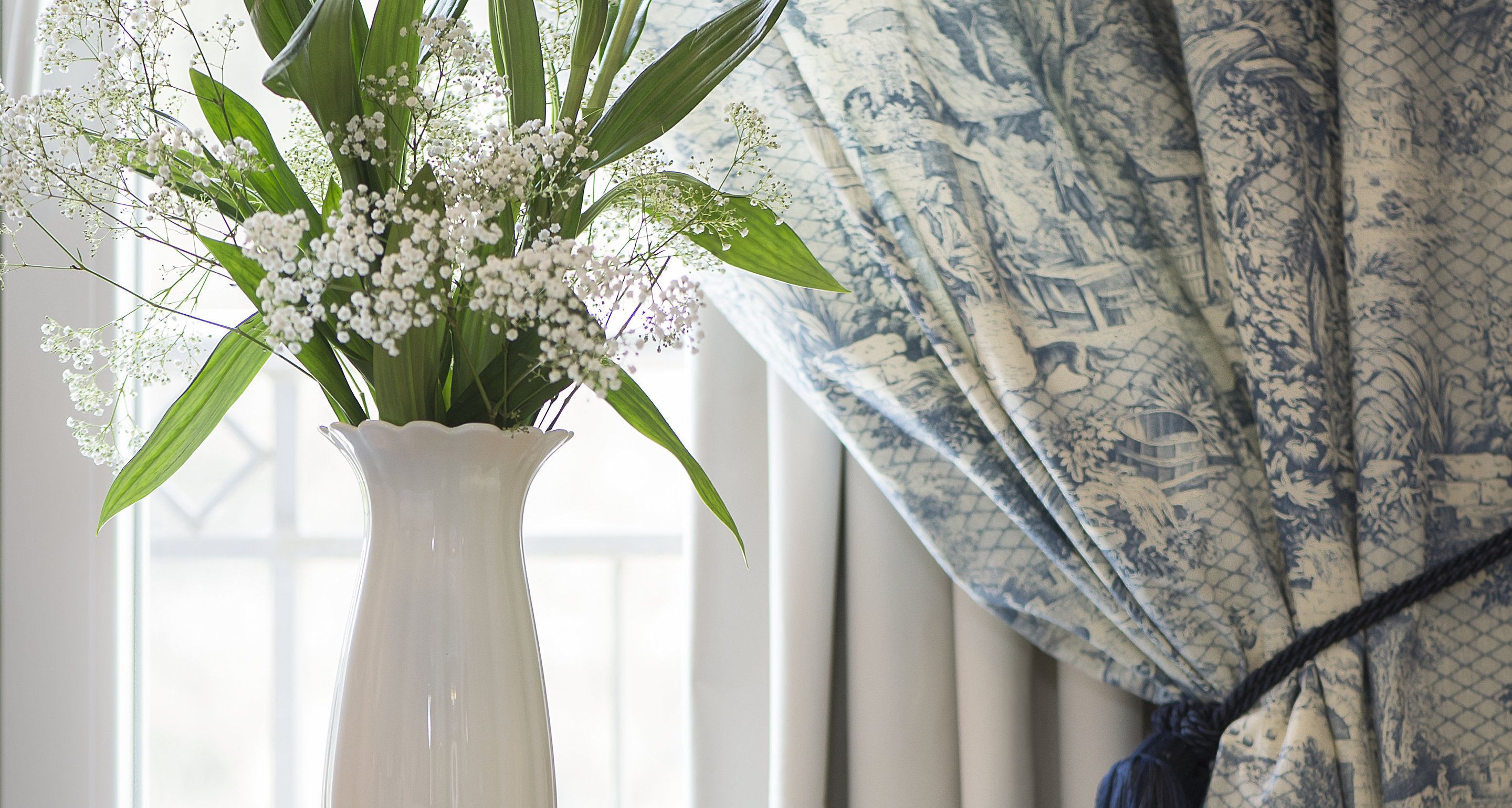 A white vase with white flowers and green leaves on a windowsill.