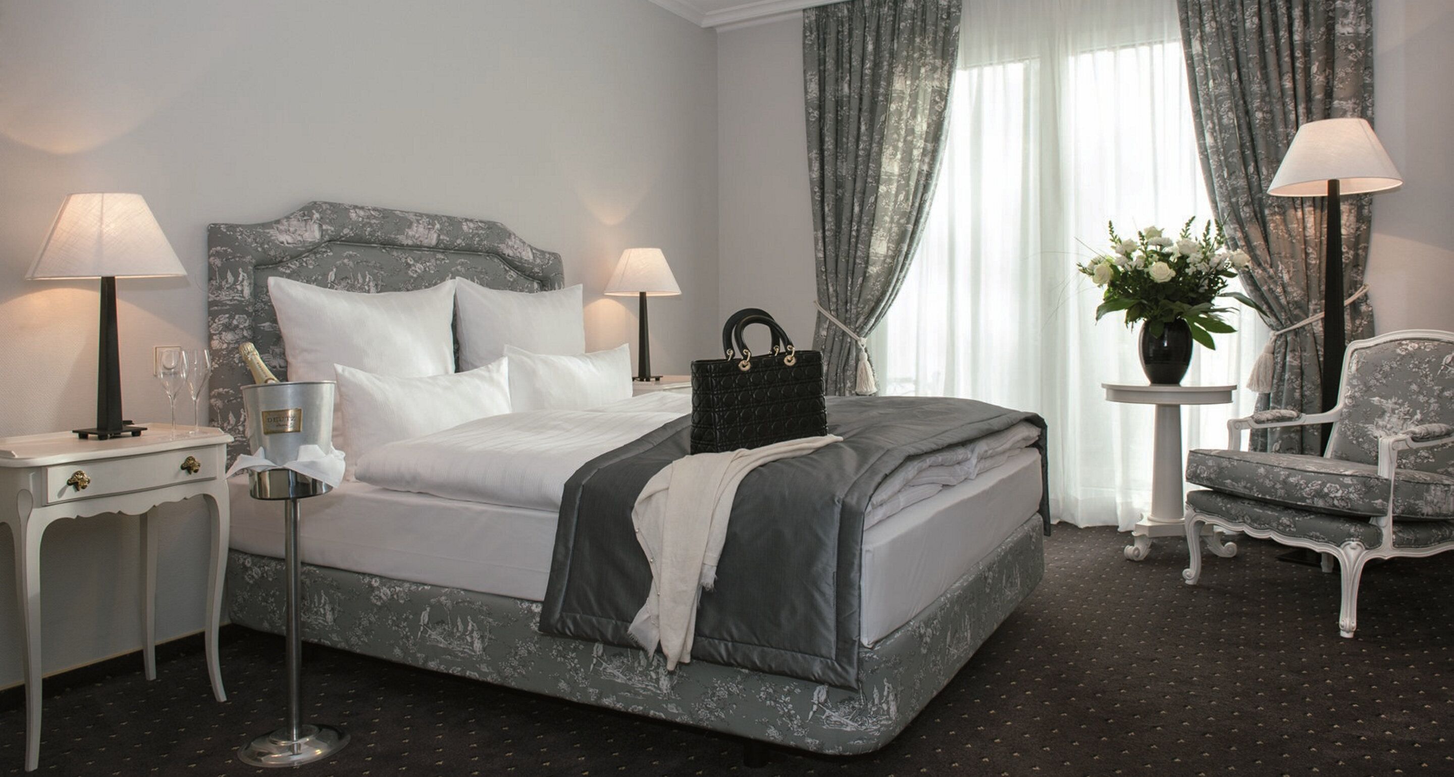 Habitación de hotel con una cama, dos mesitas de noche, una silla, una planta en maceta y una ventana con cortinas.