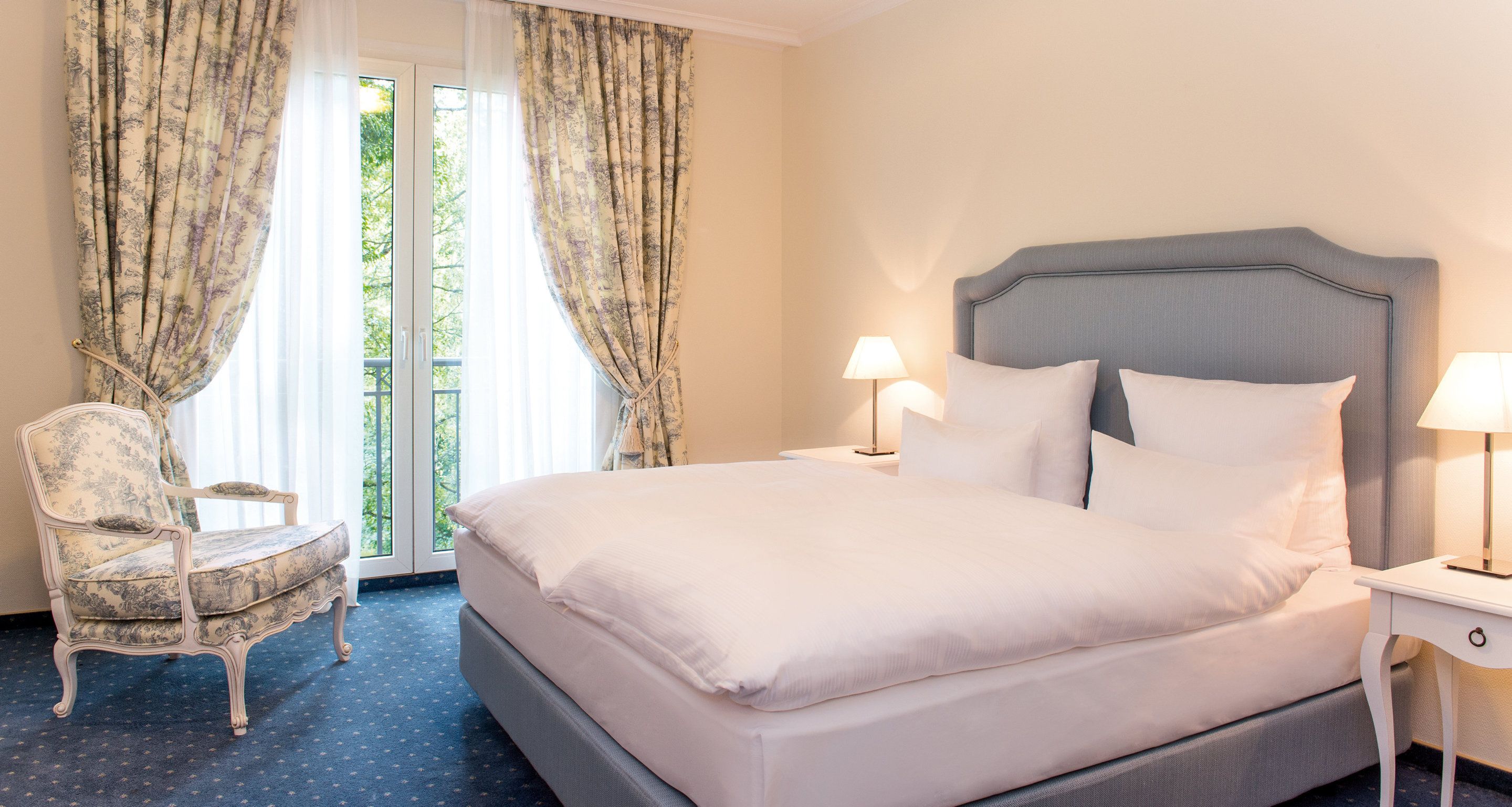A hotel room with a large bed, two bedside lamps, a chair, and a view of greenery through the window.