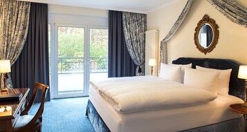 Habitación de hotel con una cama grande, un escritorio y una silla. Cuenta con un balcón con cortinas y vistas a árboles.