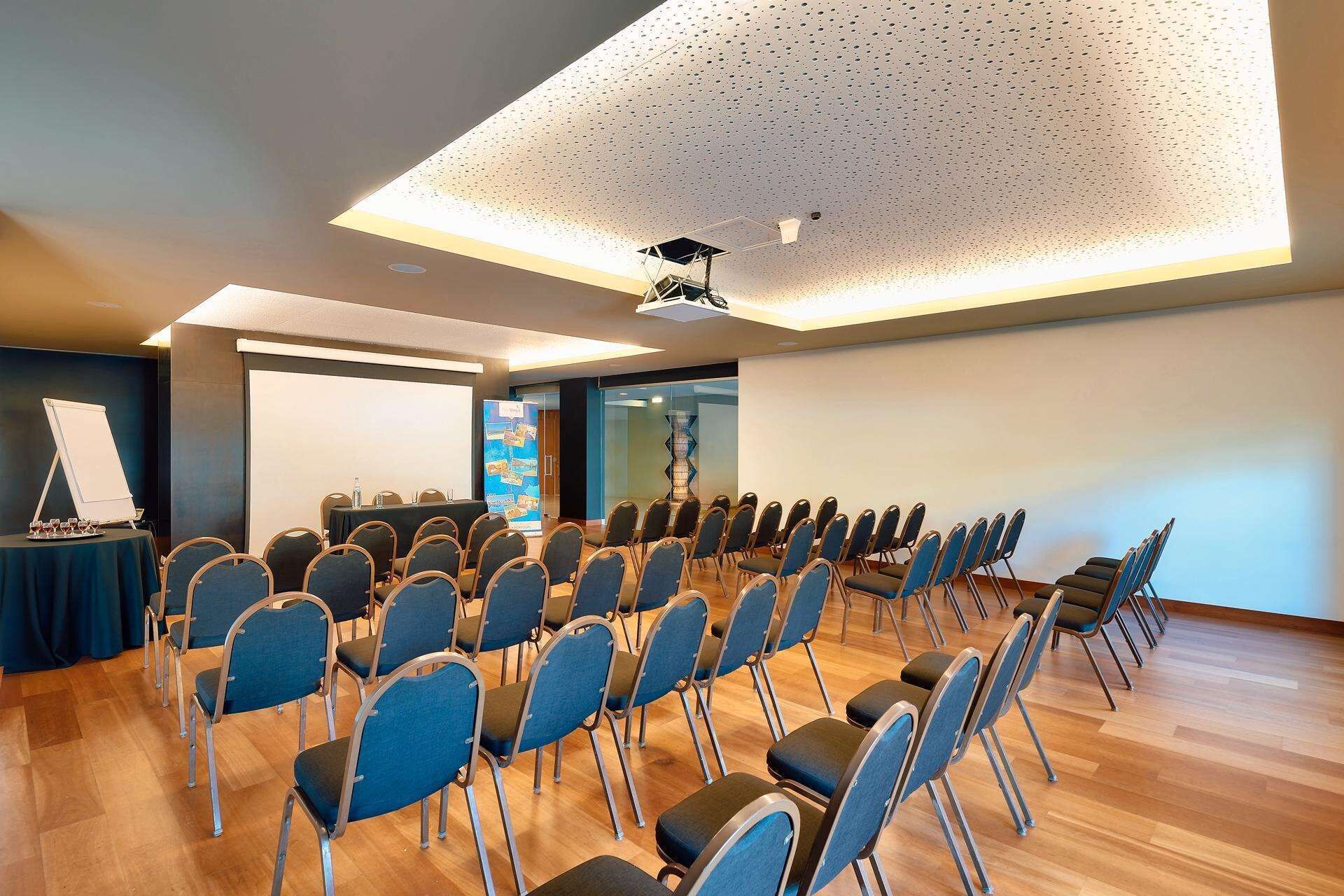 Ein Konferenzraum mit Reihen von Stühlen, einer Projektionsleinwand und einer Whiteboard-Tafel.