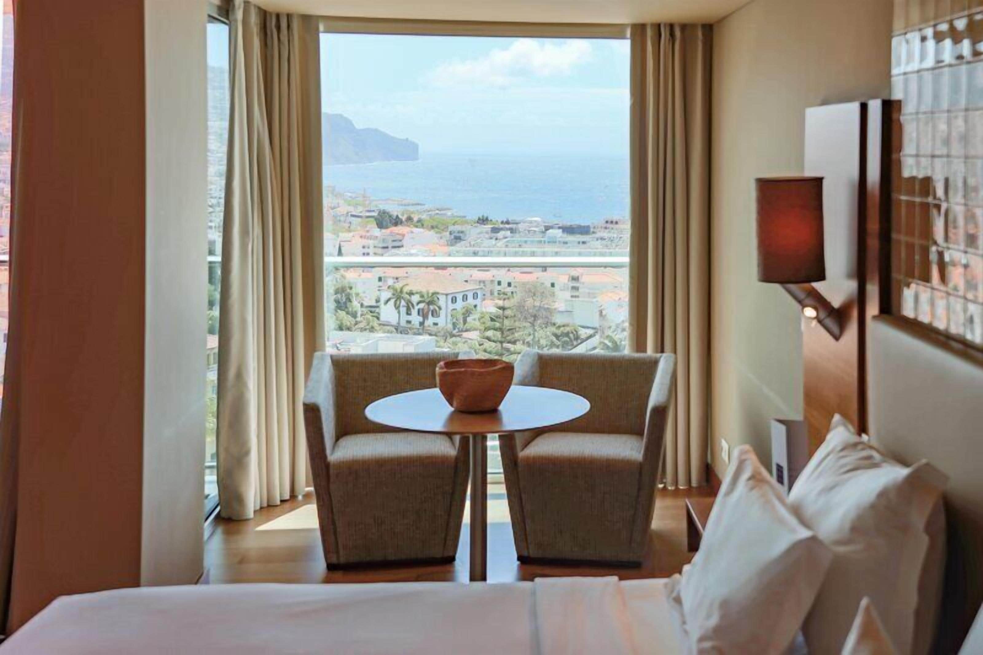 A hotel room with a bed, two armchairs, a small table, a view of the sea and buildings, and a wall-mounted lamp.