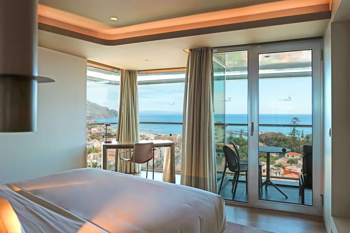 Une chambre d’hôtel avec un grand lit, une petite table, deux chaises et un balcon offrant une vue sur la mer et les bâtiments.