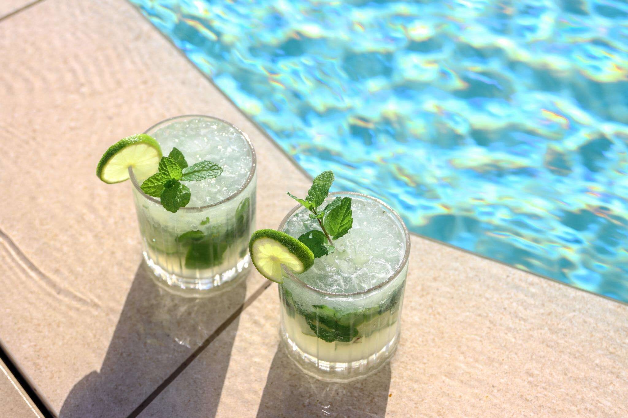 Two glasses of a beverage with lime and mint garnish, placed on a stone surface by a pool.