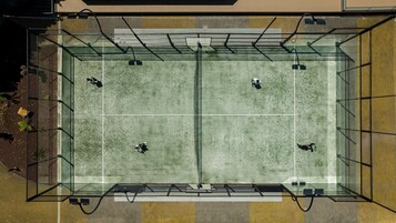 A tennis court with four players, surrounded by a glass enclosure, and a brick wall on one side.