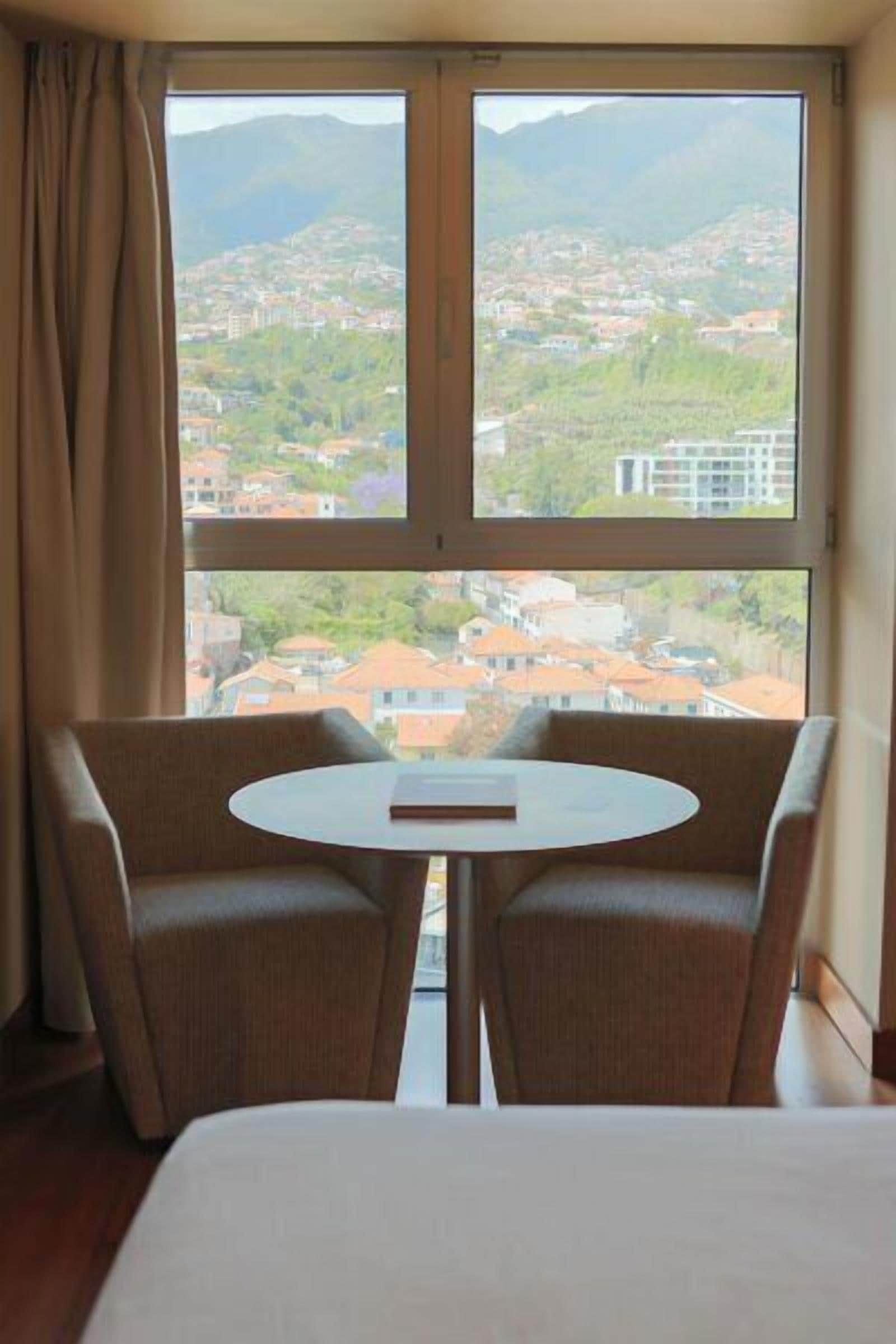 A hotel room with a bed, a table, two chairs, and a large window with a view of a town and mountains.