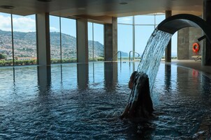 Ein großes Hallenbad mit Wasserfall, großen Fenstern und Blick auf eine hügelige Landschaft.