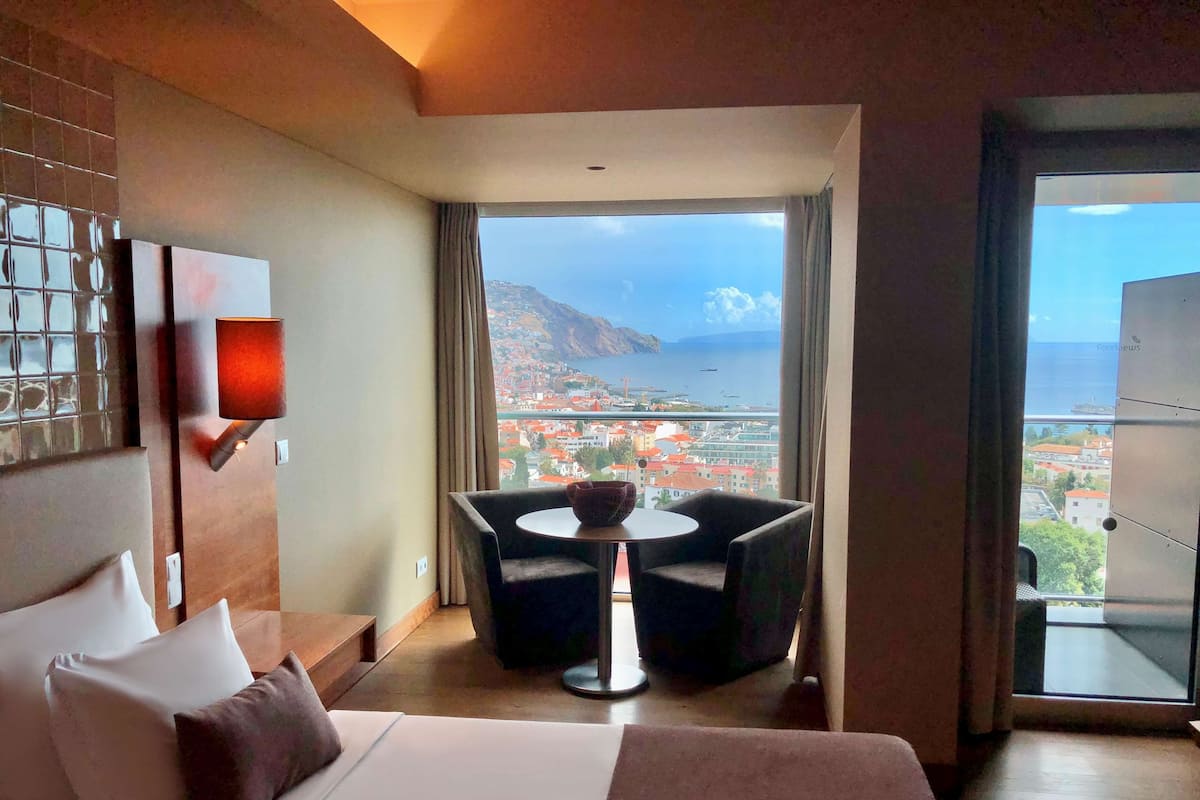 Une chambre d’hôtel avec un grand lit, une petite table avec deux chaises, une vue sur une ville côtière et un balcon avec une porte vitrée.