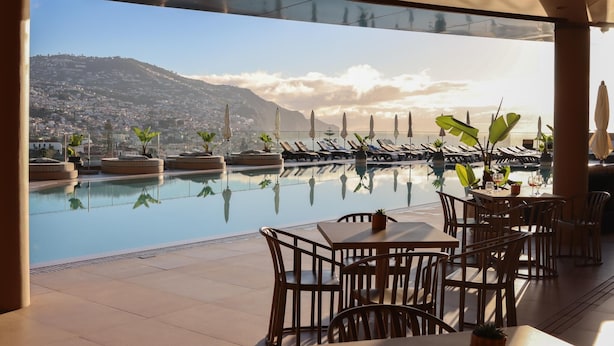 A poolside area with lounge chairs, a table with chairs, and a view of a mountain and cityscape.