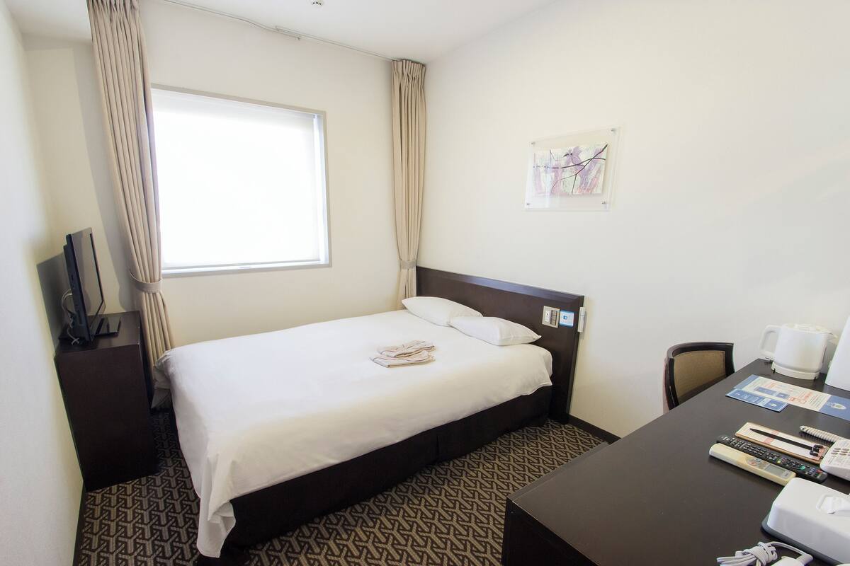 A hotel room with a bed, a desk with a chair, a window, and a wall-mounted artwork.