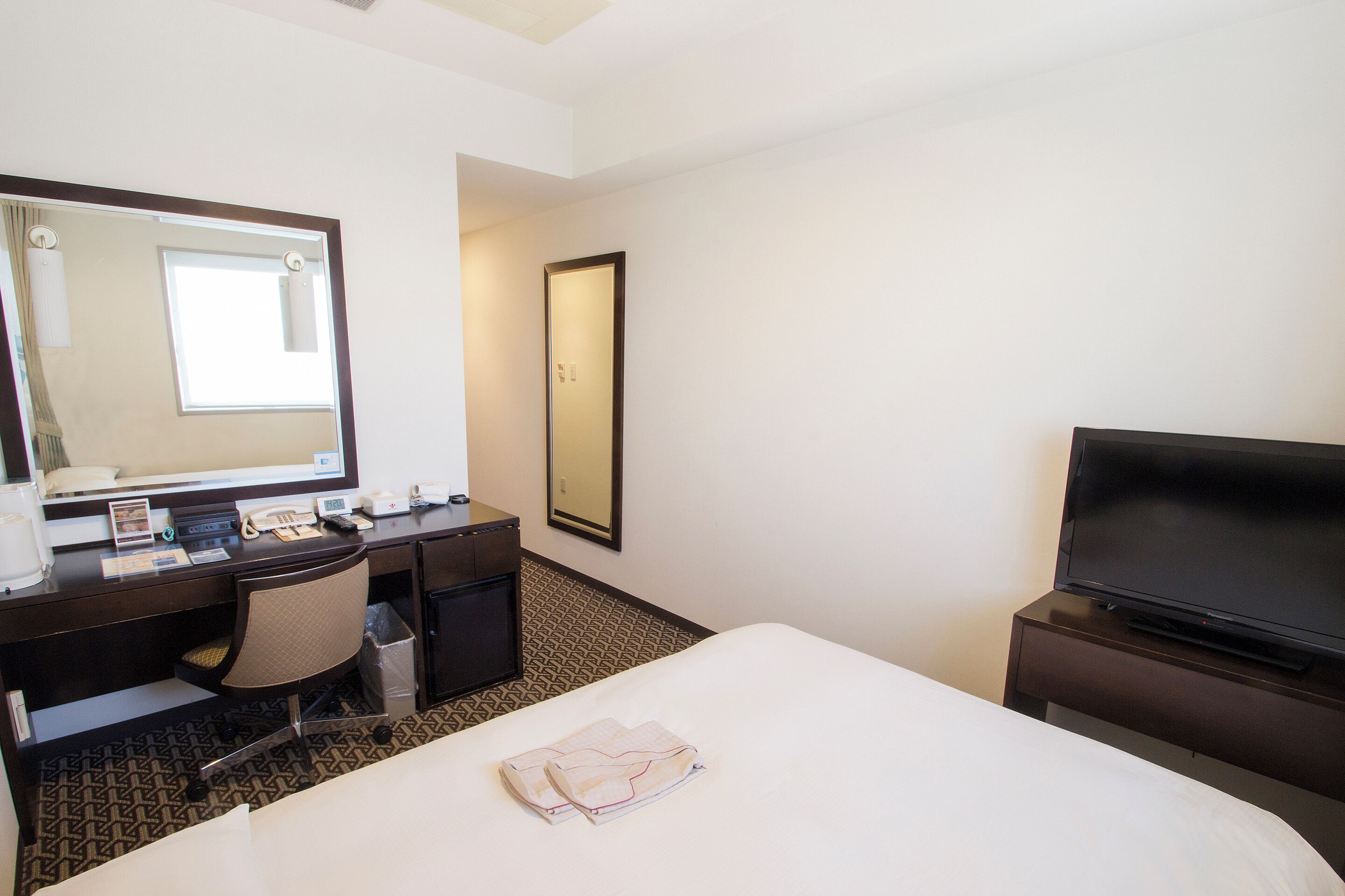 Une chambre d’hôtel avec un lit, un bureau équipé d’un ordinateur, une télévision et un grand miroir.