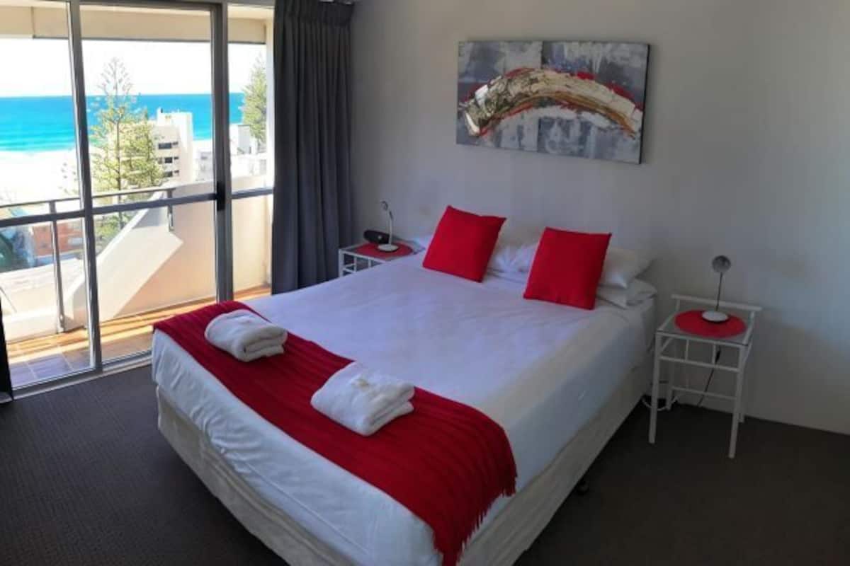 A hotel room with a large bed, red pillows, a painting on the wall, and a view of the ocean.