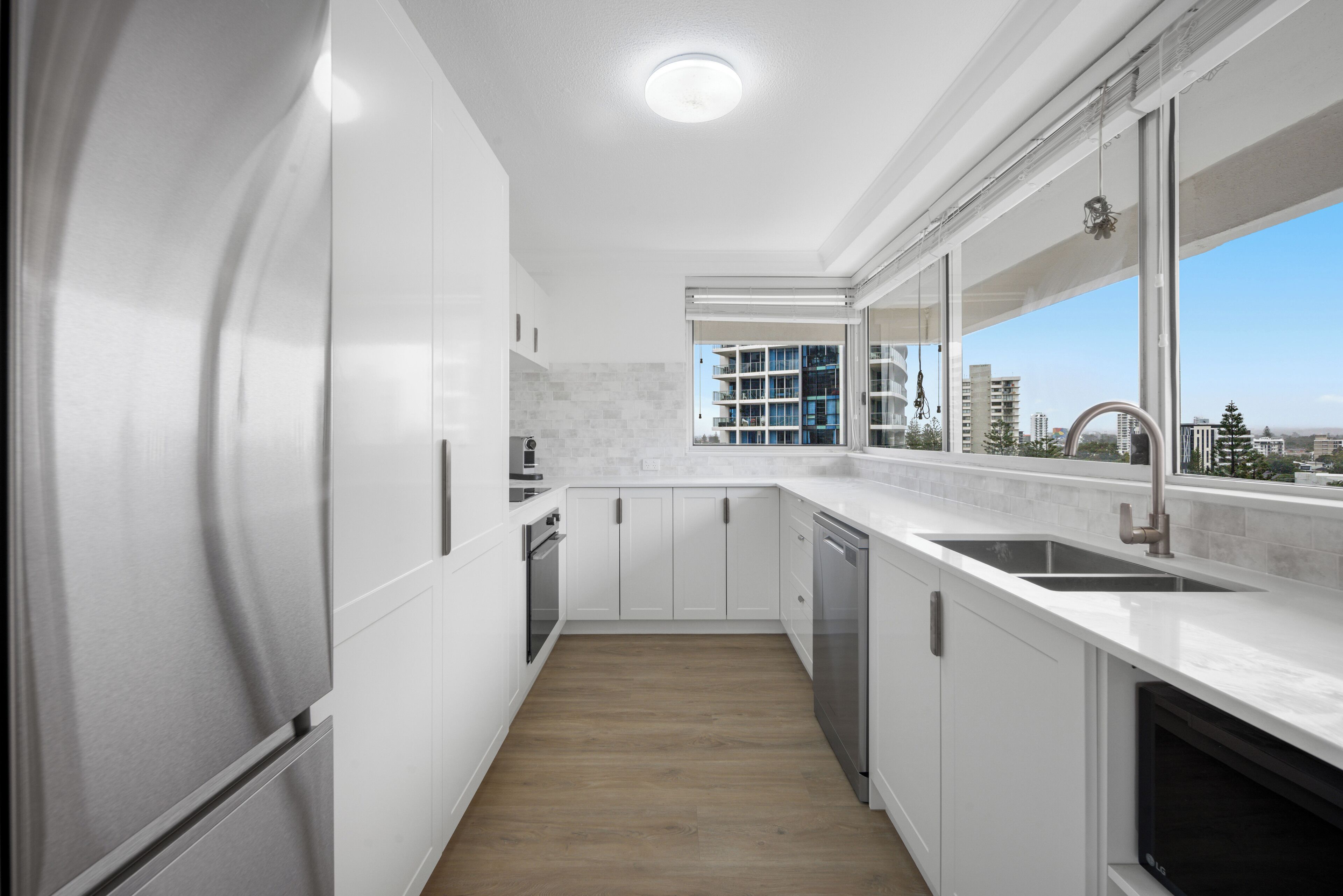 Een moderne keuken met witte kasten, een houten vloer, een groot raam met uitzicht over de stad en een inbouwvaatwasser.